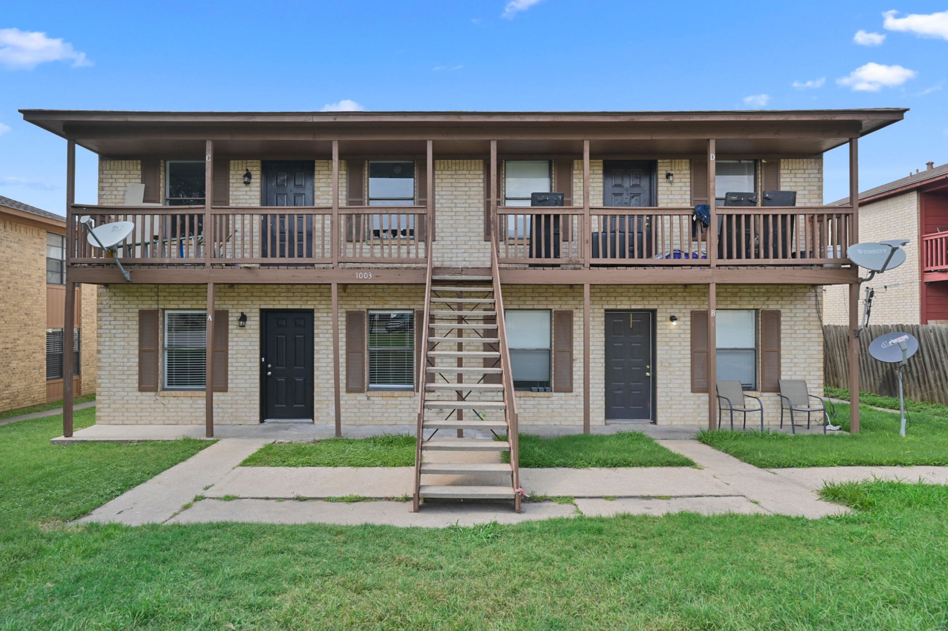 Two-story, beige brick apartment building with brown wood railings, stairs, and dark doors under a blue sky.