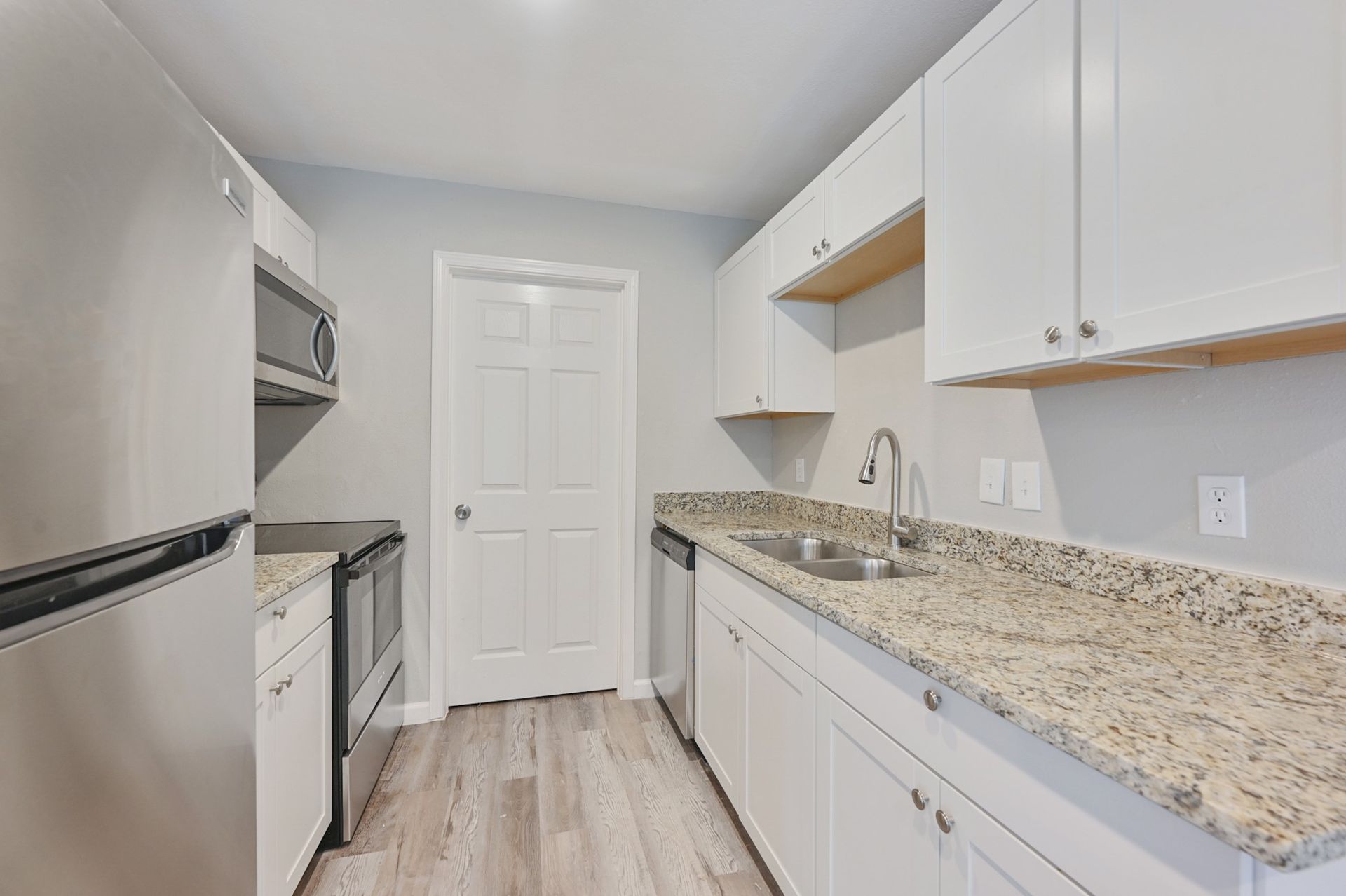 A compact kitchen with white cabinets, speckled granite countertops, stainless steel appliances, and light wood flooring.