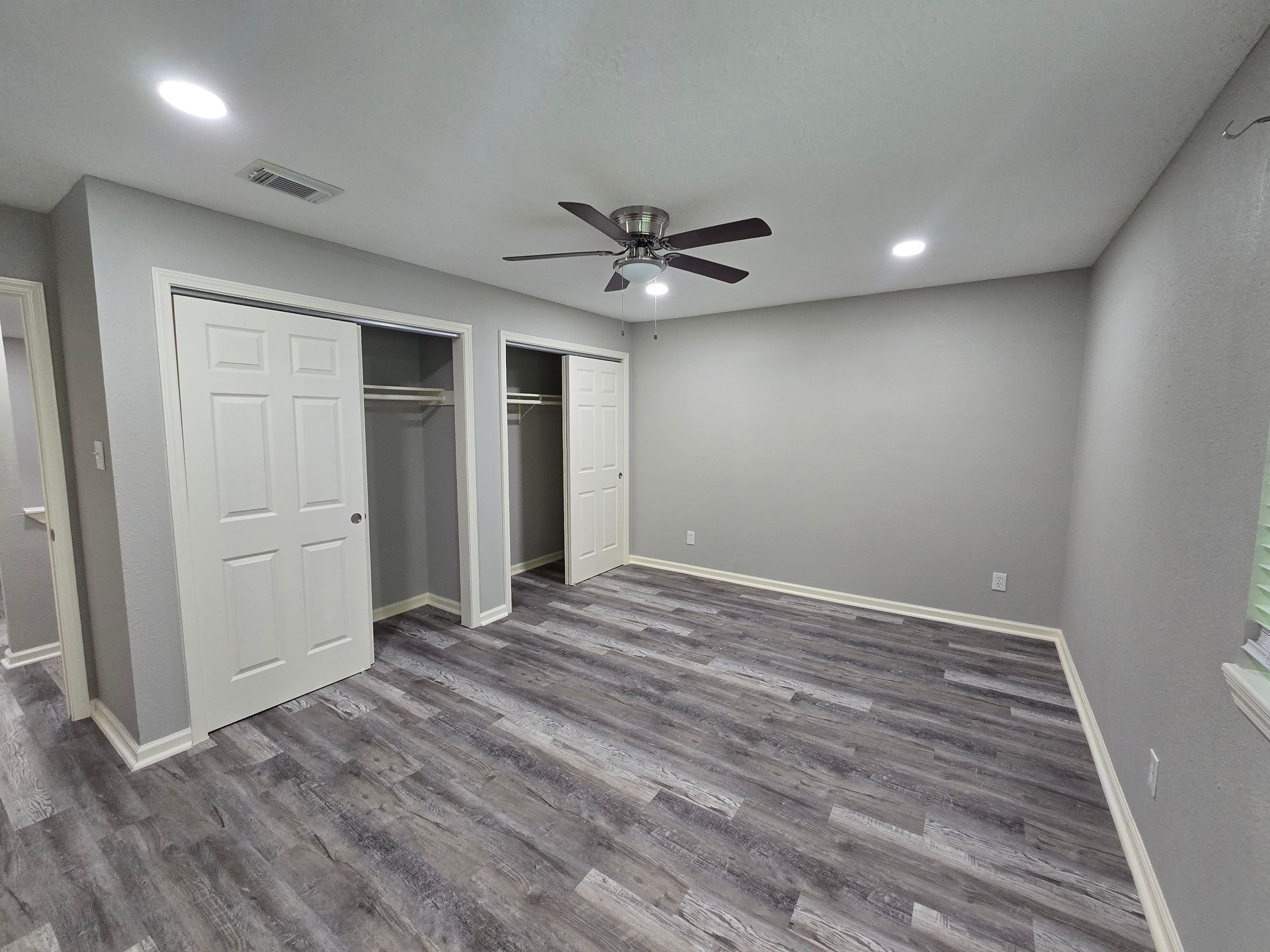 An empty bedroom with gray walls, wood-look flooring, two open white closets, a ceiling fan, and recessed lighting.