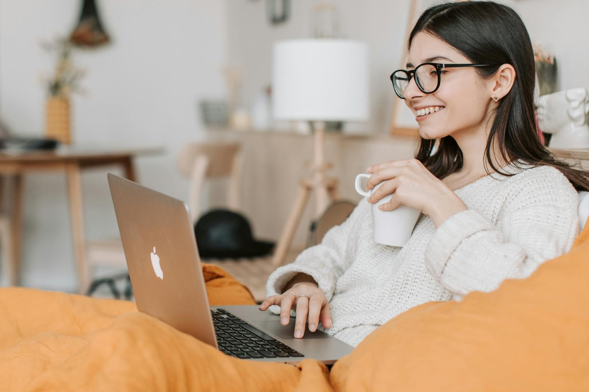 Woman with glasses using a laptop, holding a mug, and smiling on a sofa.