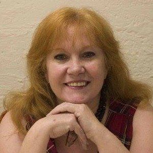 A woman with red hair is smiling with her hands folded in front of her face.