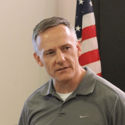 A man in a grey nike shirt is standing in front of an american flag.
