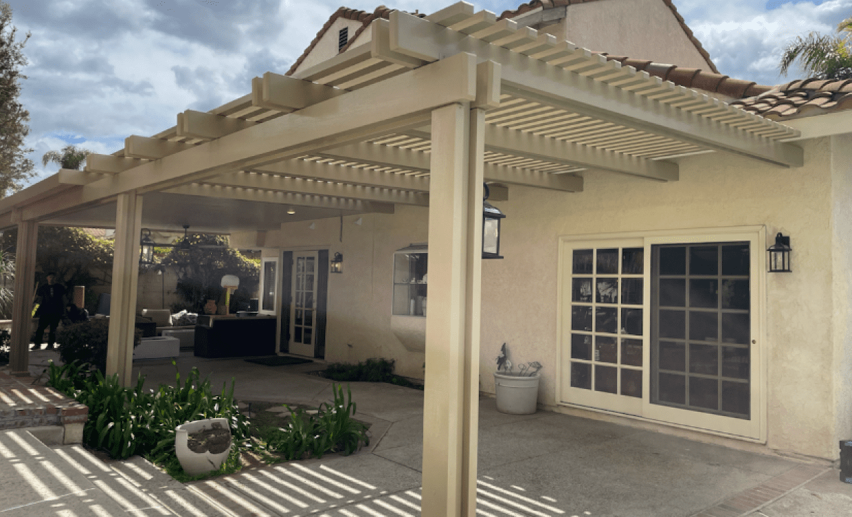 A pergola covers a patio attached to a house with sliding glass doors. Beige structure, sunny day.