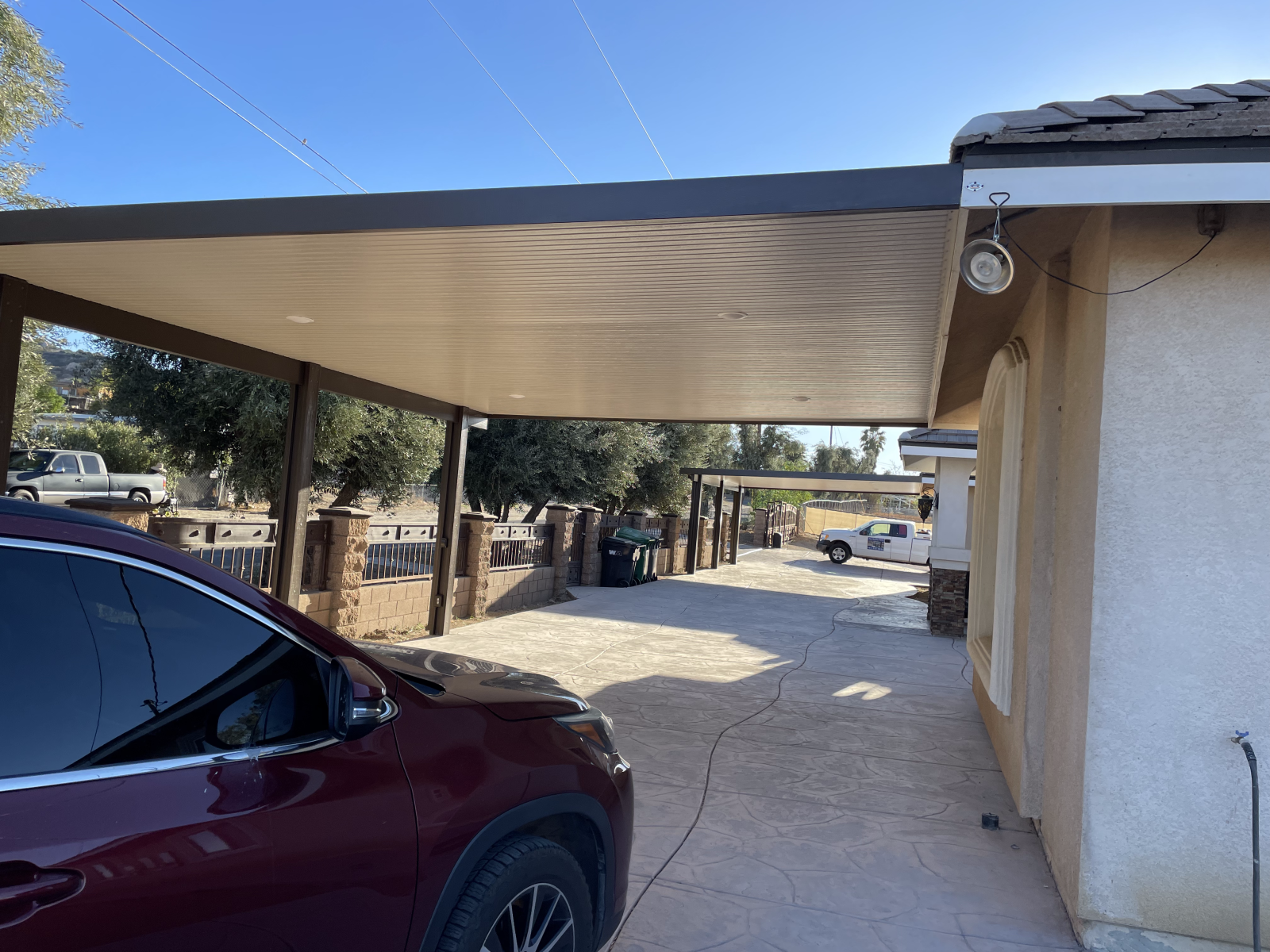 Carport with a parked maroon car and a driveway next to a beige house under a clear sky.