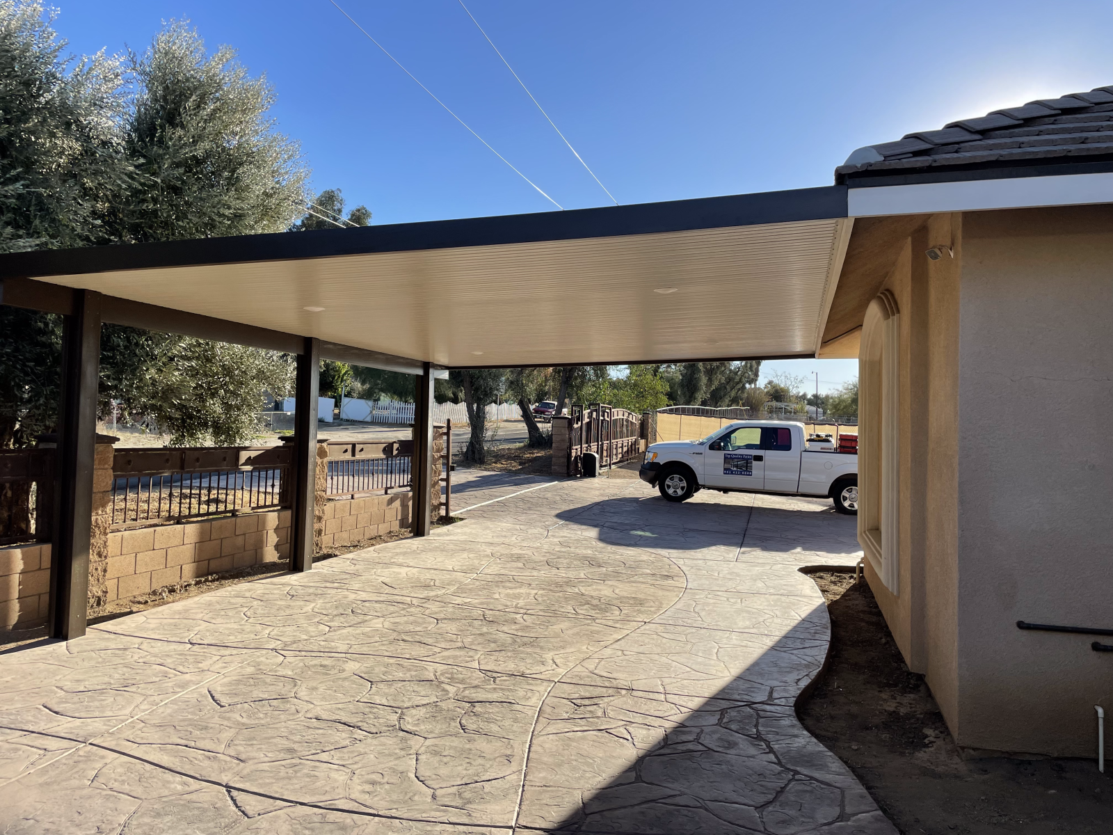 Carport with white truck parked beneath, concrete driveway, attached to a tan building.