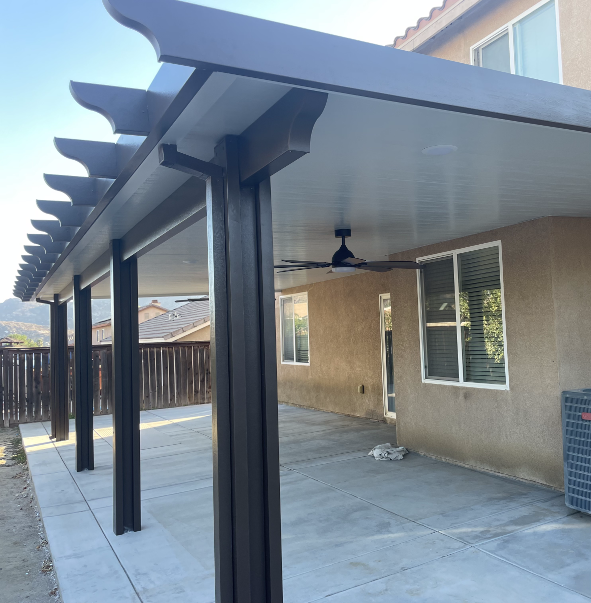 Patio cover with dark brown supports and gray concrete floor, attached to a light brown stucco house.