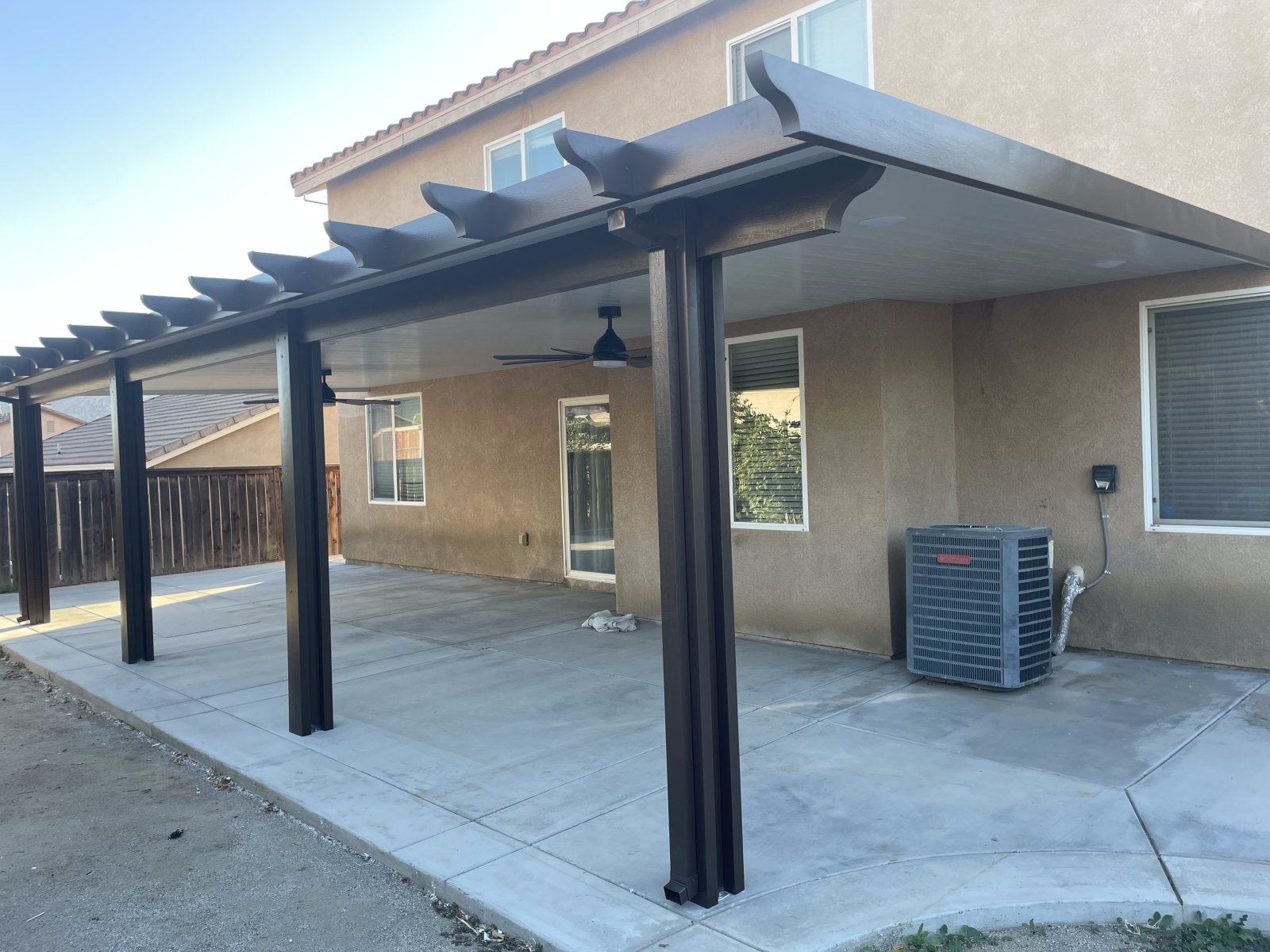 Patio cover attached to a two-story beige house with dark columns and a ceiling fan.