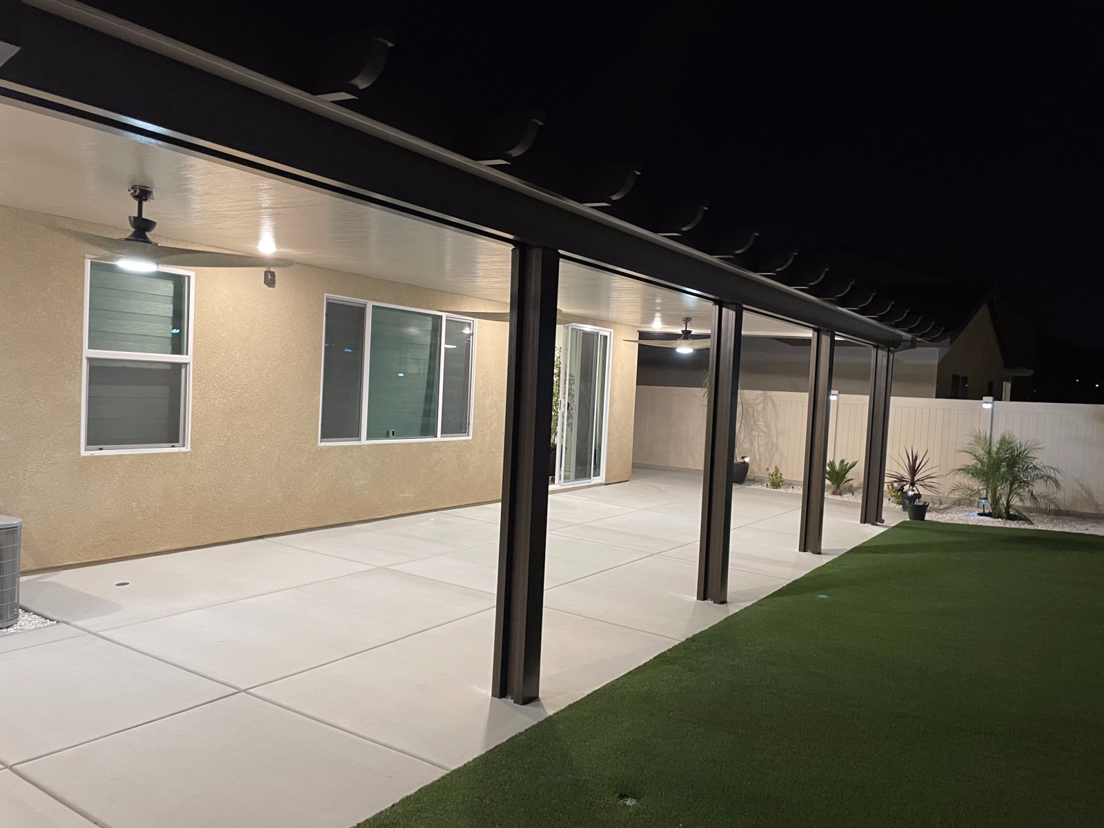 Patio with dark-colored posts and roof. Concrete flooring, artificial grass, and house in the background at night.