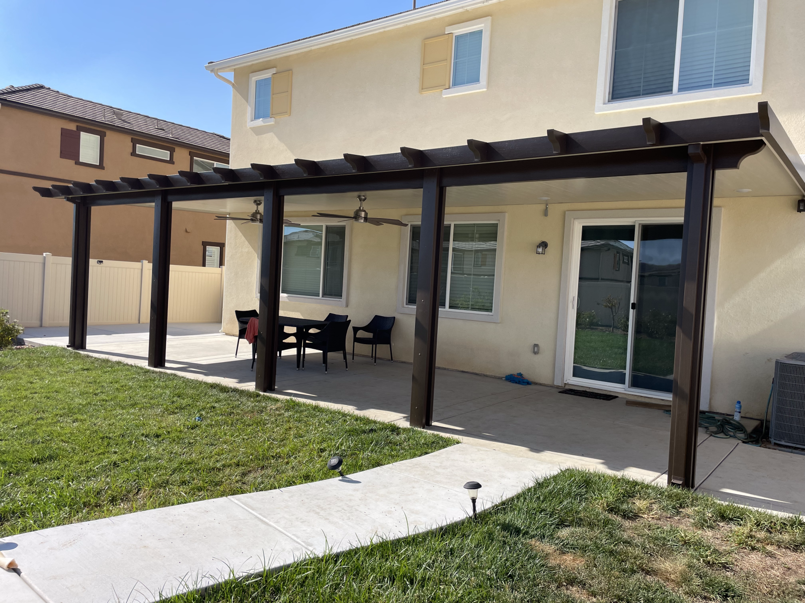 Backyard patio with brown pergola, concrete pathway, and lawn.