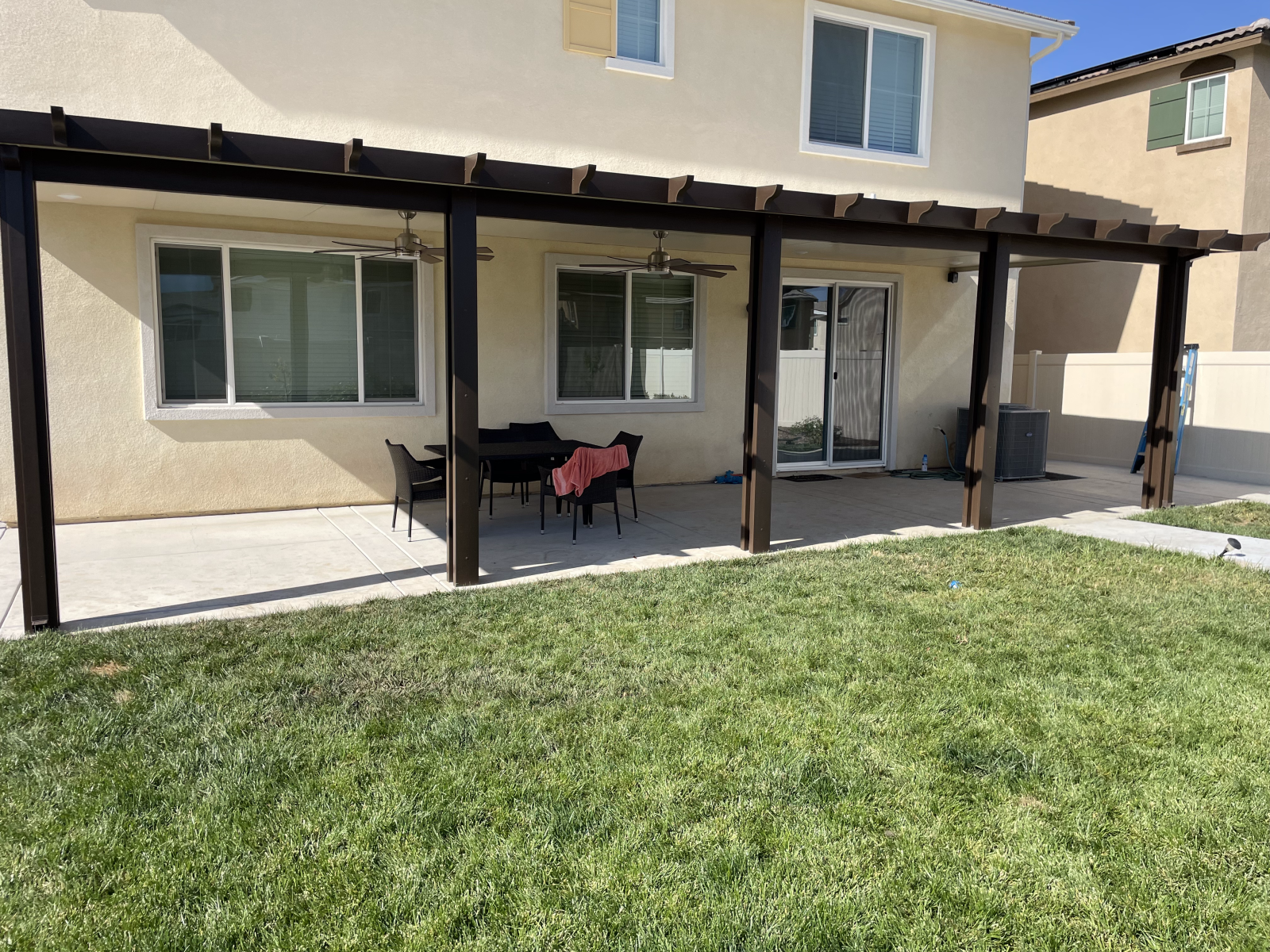 Backyard patio with pergola, table, and chairs. Green grass and beige house in background.