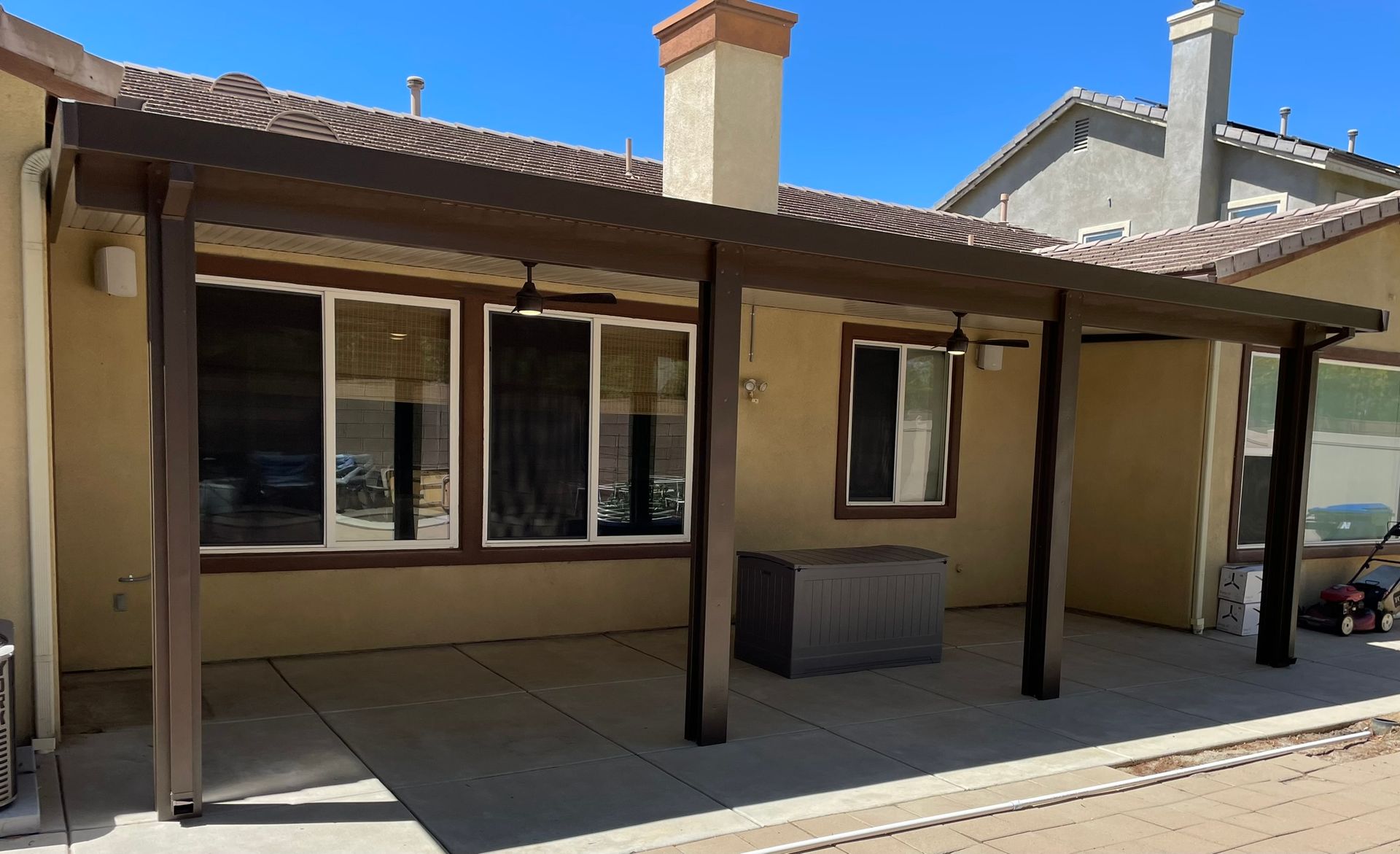 Patio with brown pergola, attached to a house with windows and a concrete floor.