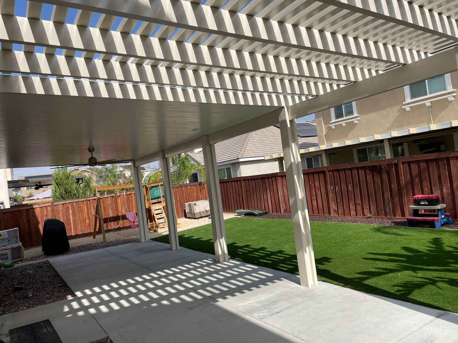 Covered patio with white beams and concrete floor, overlooking a grassy backyard with a brown fence.