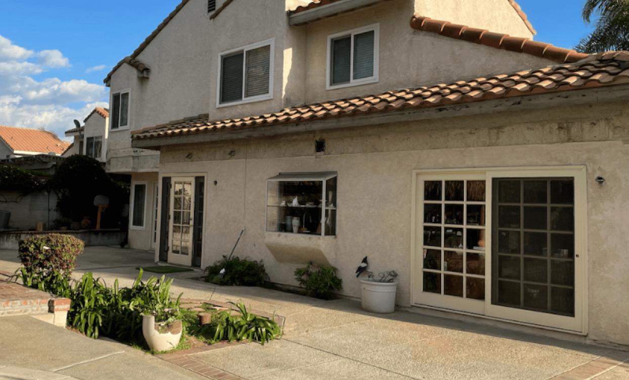 Two-story beige building with windows and sliding glass doors. Red tile roof, concrete patio, and plants in foreground.