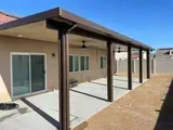 Brown patio cover with columns extending from a beige stucco home onto a concrete patio, with a clear blue sky.
