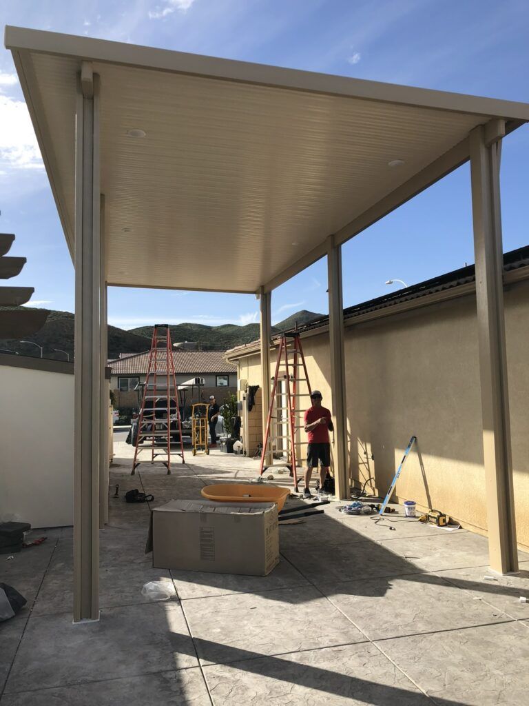 Tan patio cover construction in progress, with workers, ladders, and tools visible.