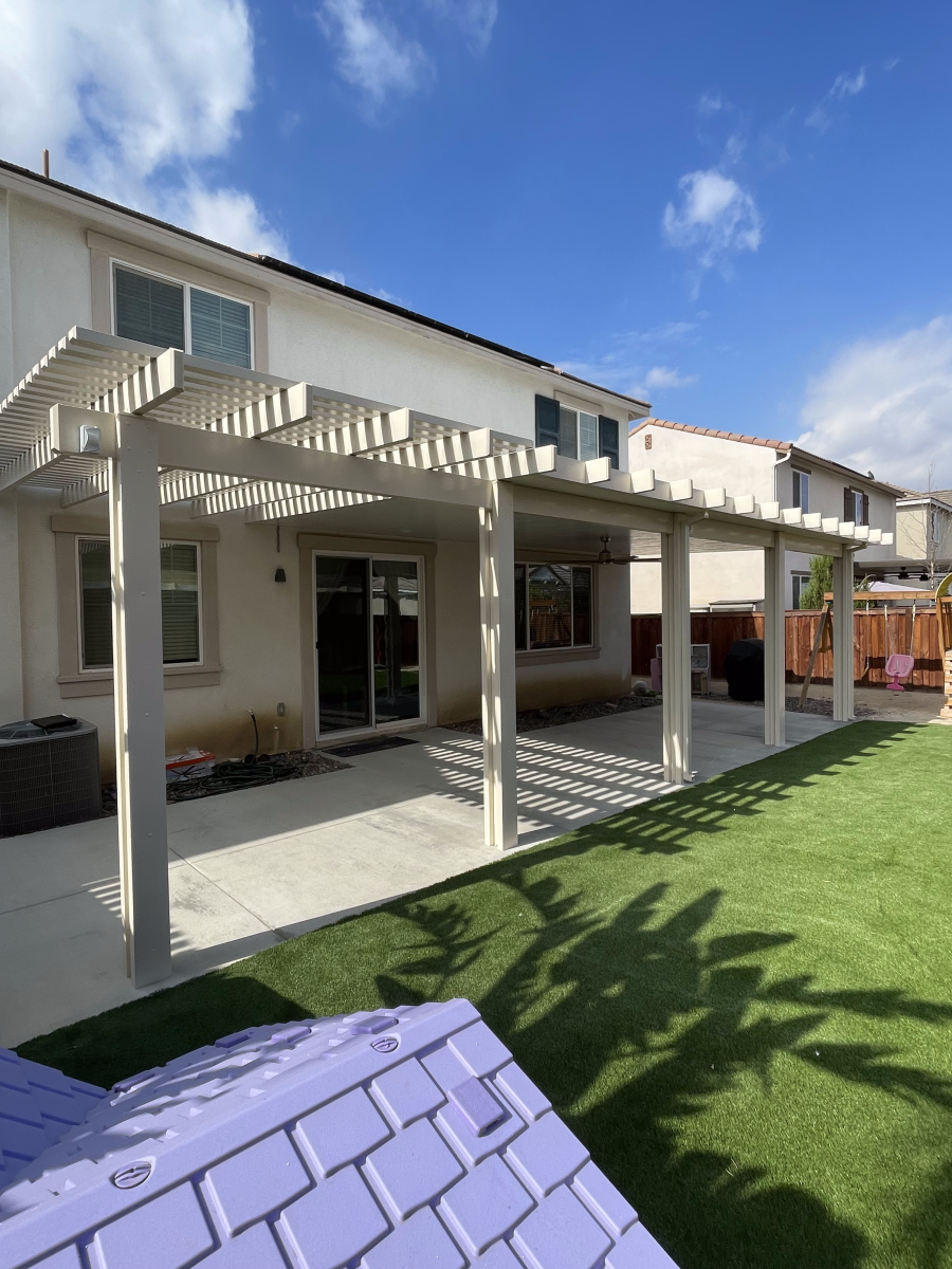 Backyard with patio and pergola over concrete, house in background, green grass, blue sky.
