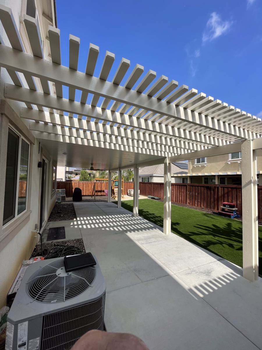 Pergola over concrete patio next to a house with a lawn and fence on a sunny day.
