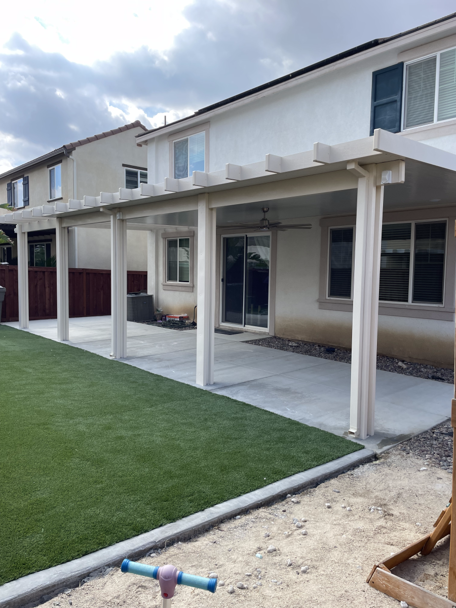 Backyard patio with white pergola, concrete floor, artificial grass, and beige house.