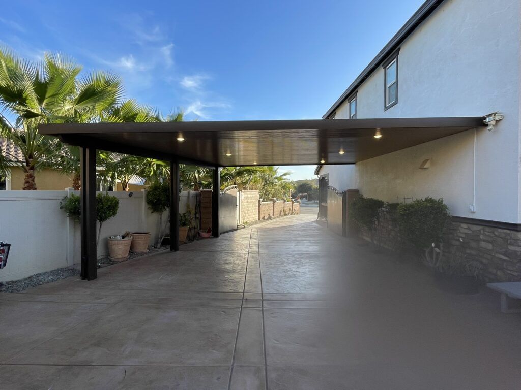 Dark brown carport attached to a two-story house, covering a concrete driveway with palm trees in the background.