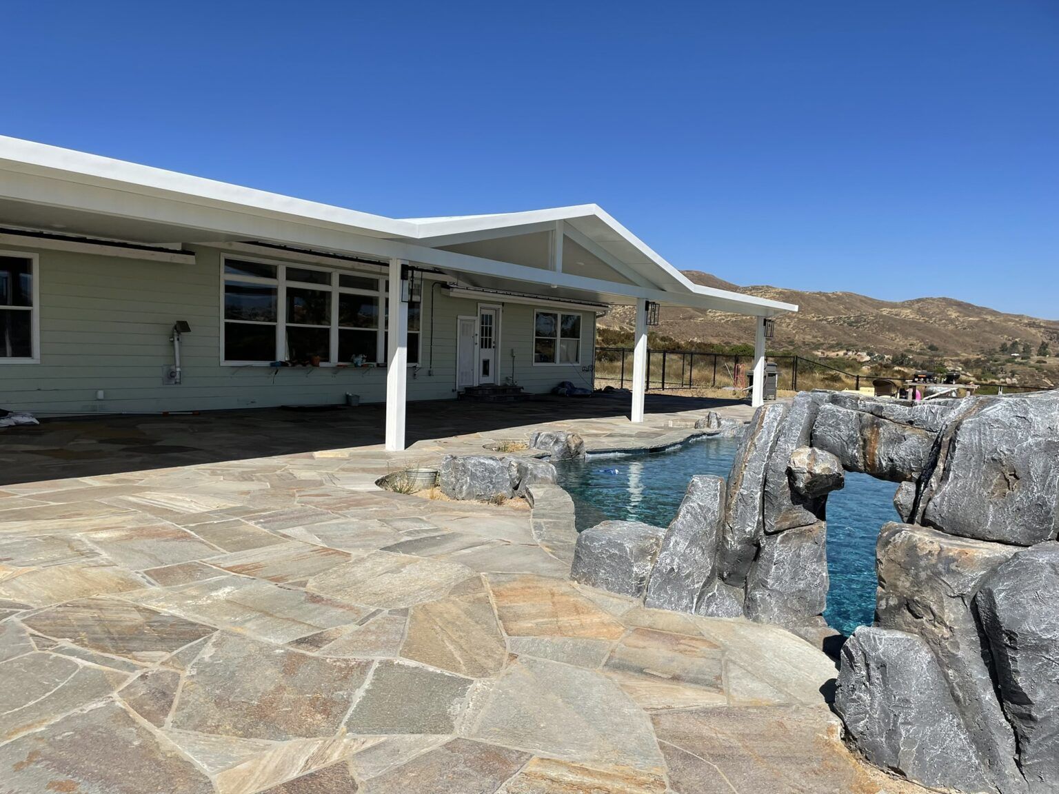 House with a stone patio and a pool surrounded by rock formations under a blue sky.