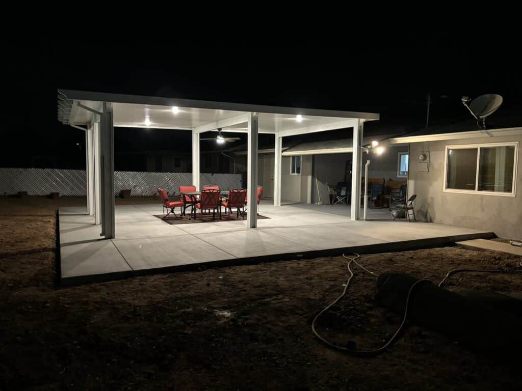 Nighttime view of a patio with a table and chairs under a lit white canopy, next to a house.