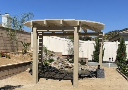 Beige pergola over a rock pond in a backyard, with a white fence and hills in the background.
