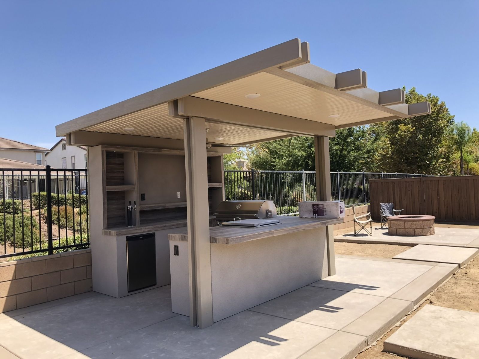 Outdoor kitchen with tan pergola, concrete counter, built-in grill, and refrigerator. Patio in backyard with fence.