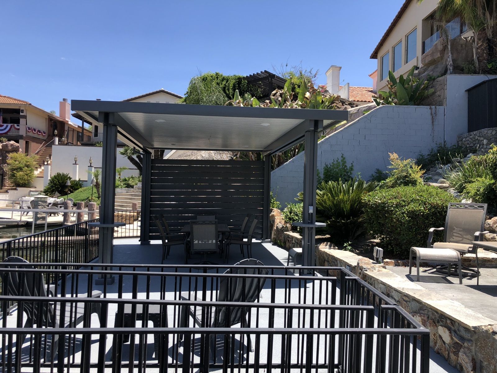 A waterfront patio with pergola and dining set. Metal railing, concrete patio, and blue sky.