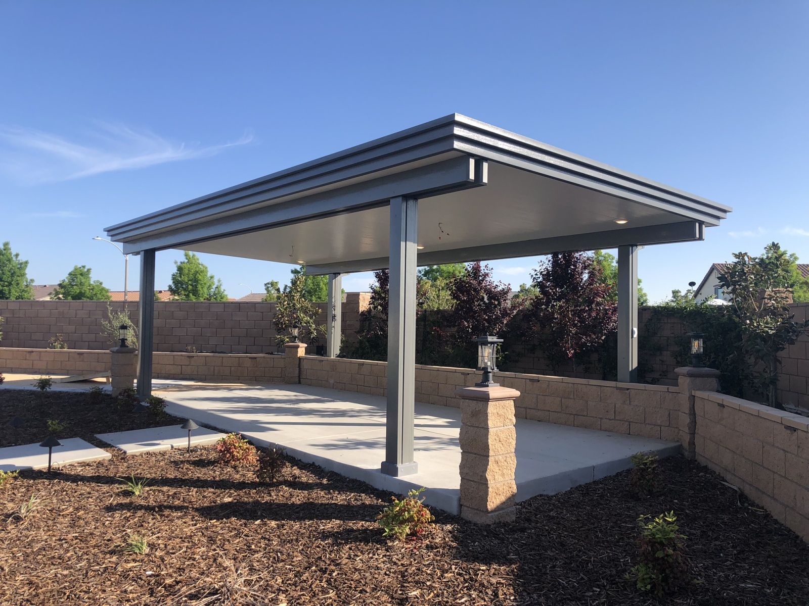 A modern silver and white patio cover with a concrete patio, surrounded by a brick wall and landscaping under a blue sky.