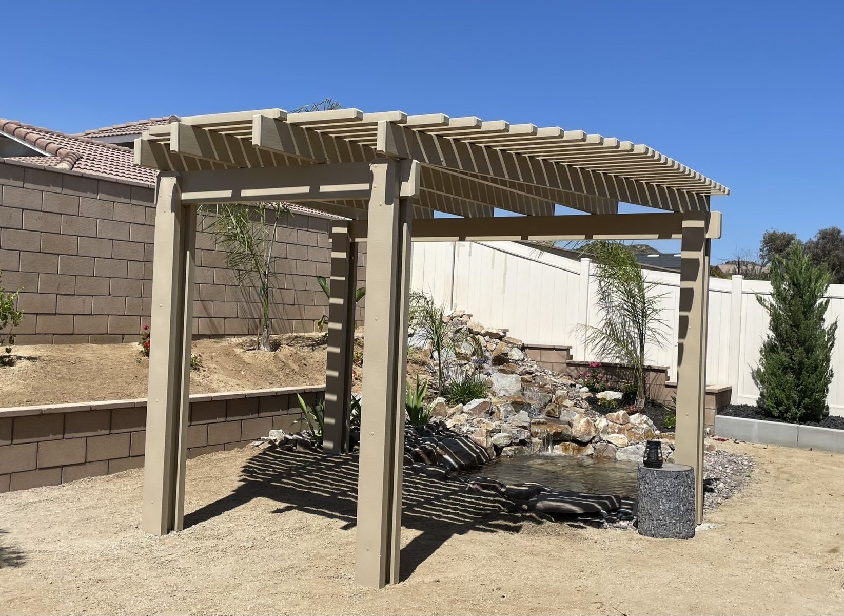 Beige pergola with curved roof over a small water feature and gravel ground, sunny day.