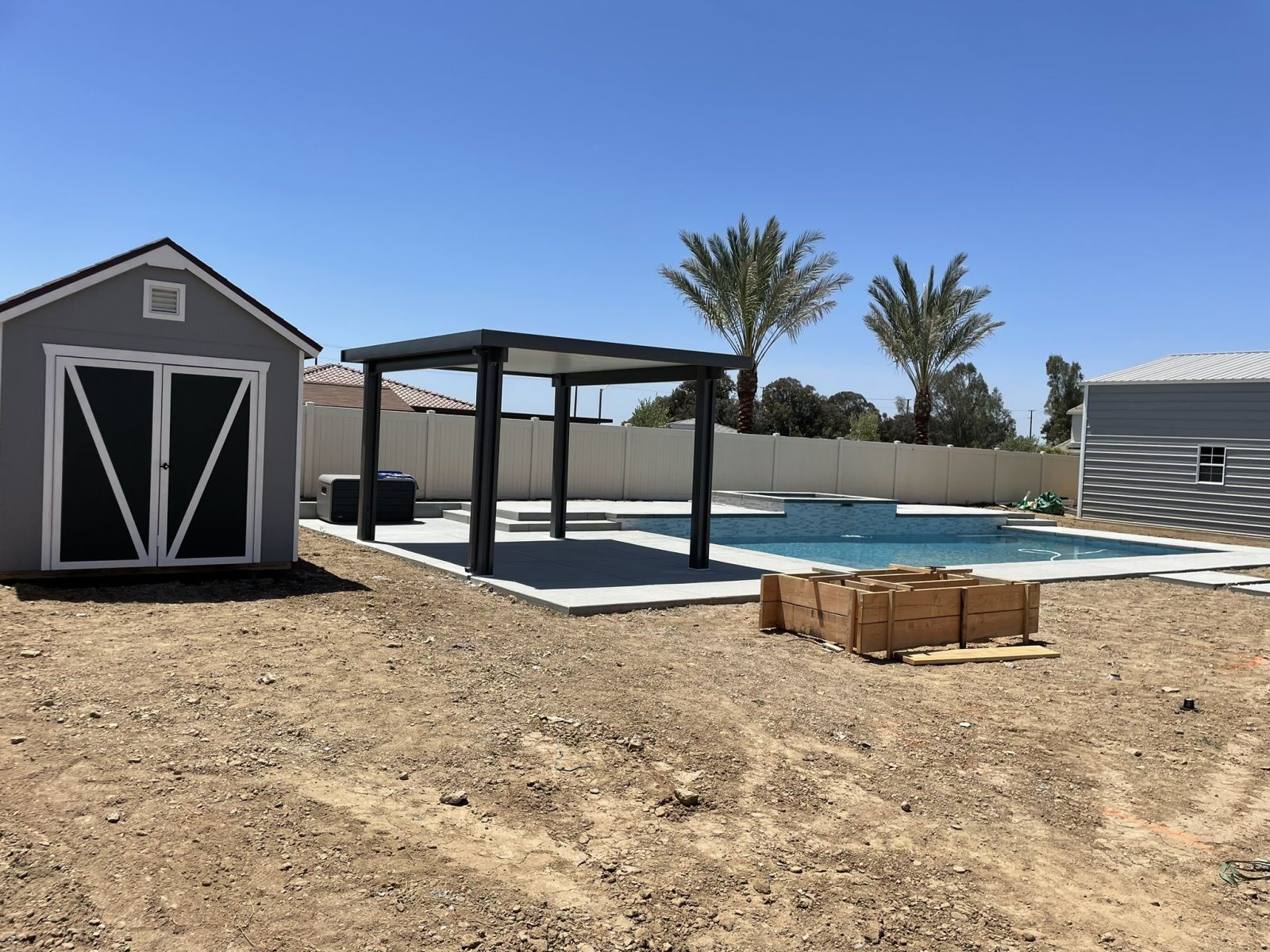 Backyard with pool, pergola, sheds, and landscaping under a blue sky.
