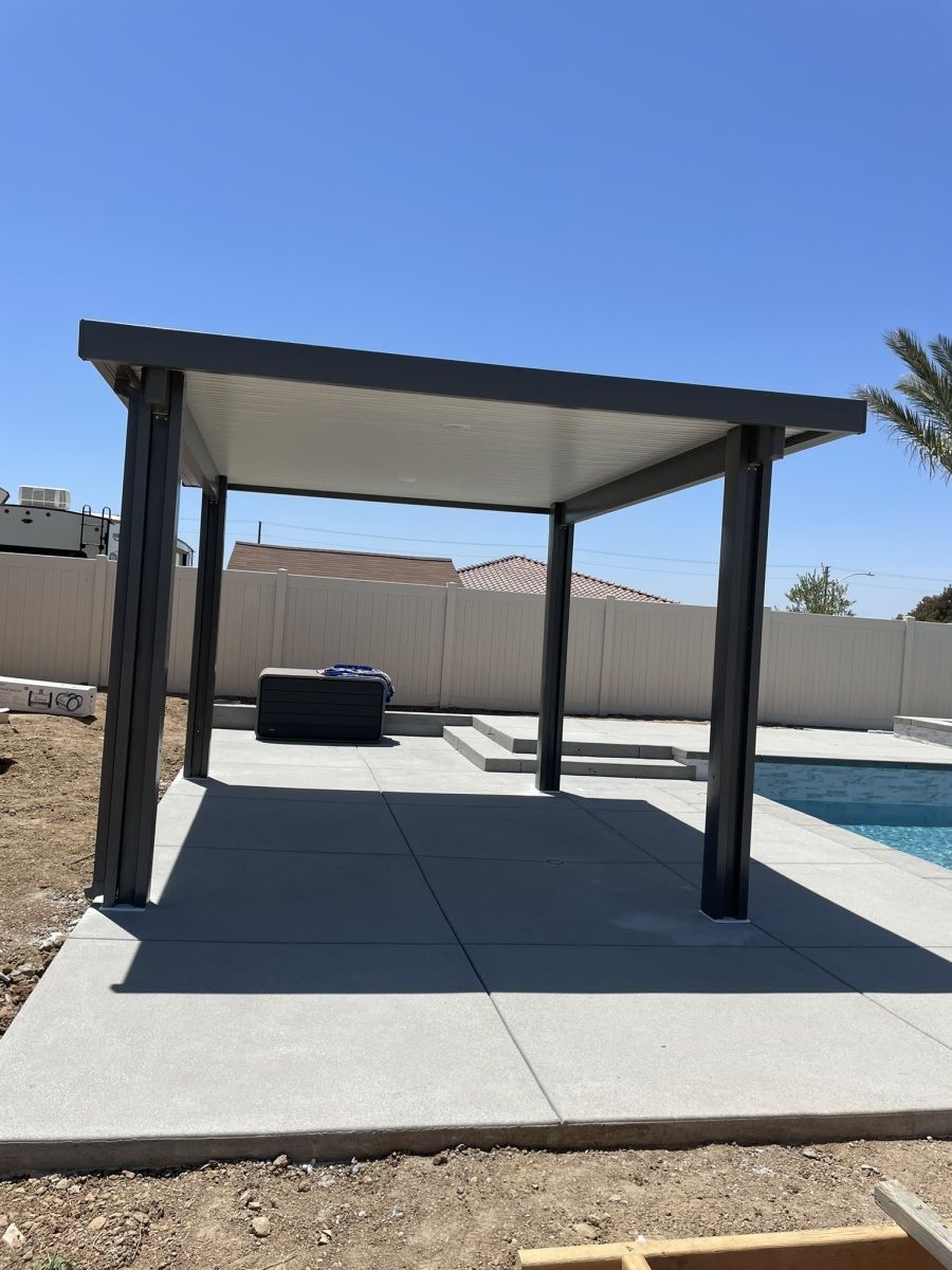 Gray patio cover over a concrete patio near a pool. Sunny day with a white fence in the background.