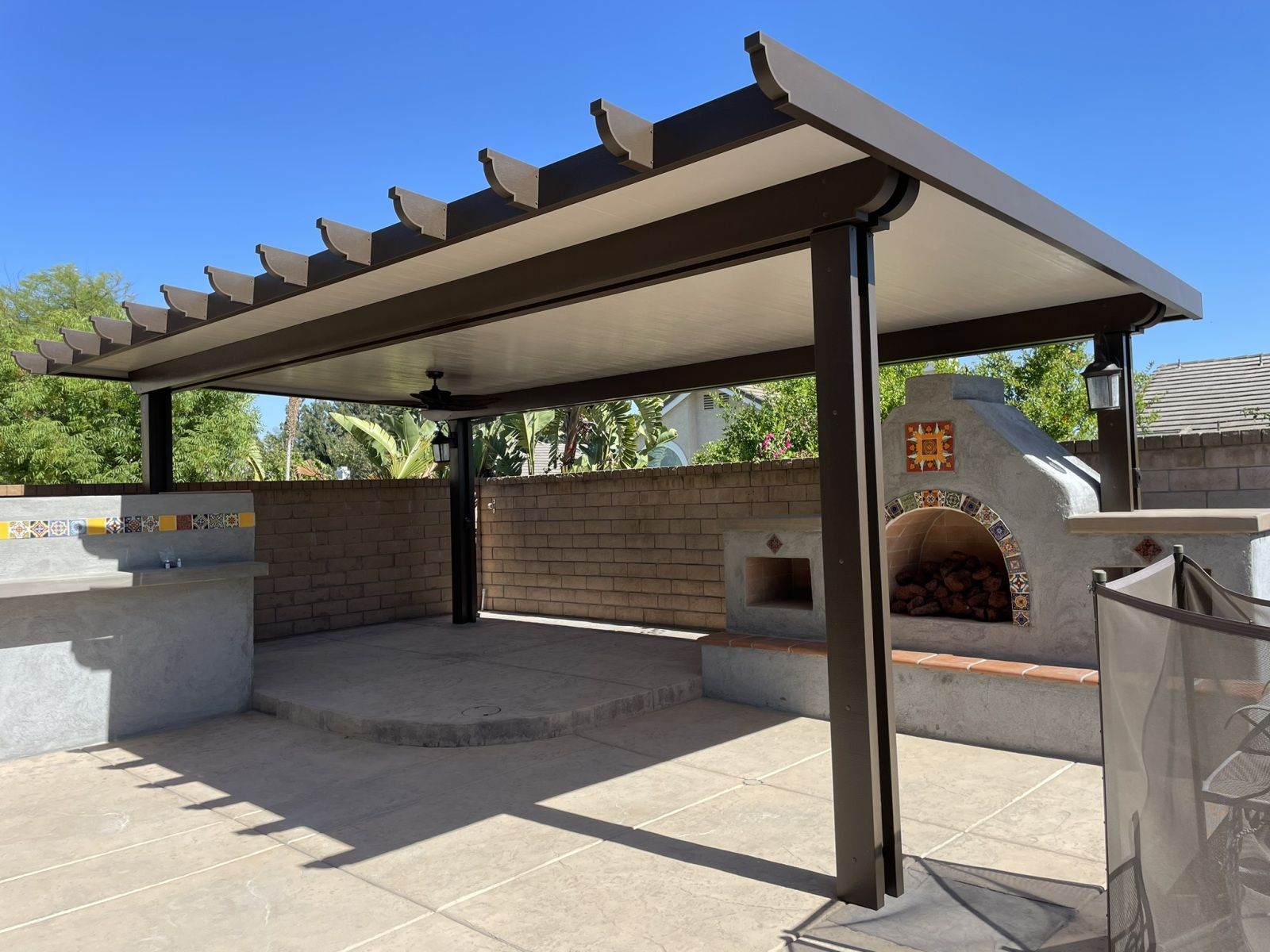 Patio with brown pergola, brick wall, outdoor fireplace, and concrete countertop under a clear blue sky.