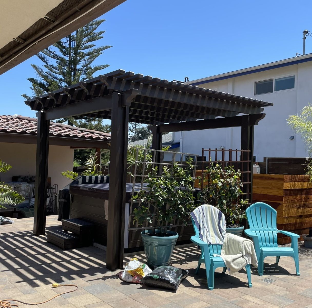 Pergola with hot tub, blue chairs, and plants in a backyard setting on a sunny day.