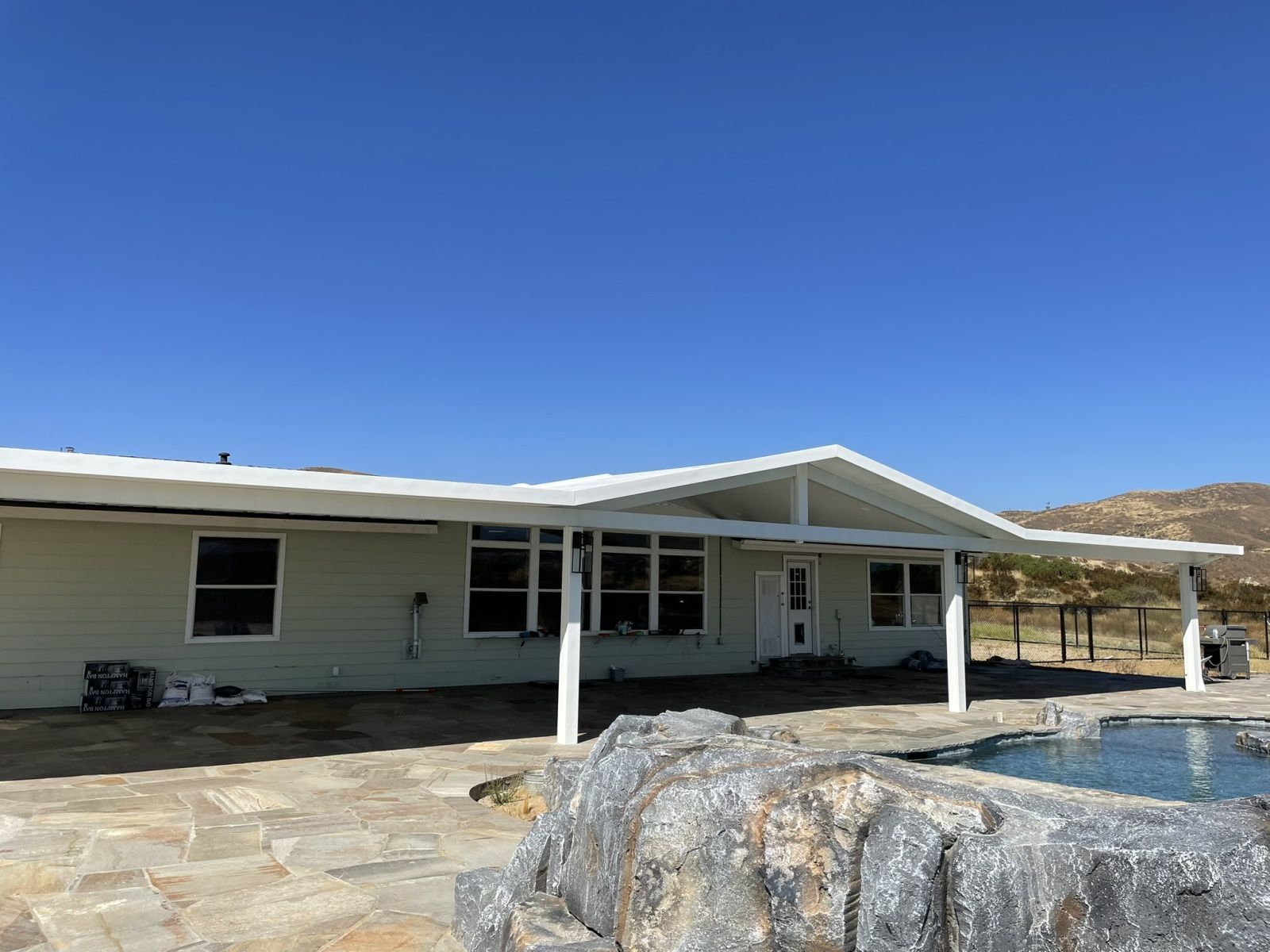 A light green house with a white patio cover, a blue pool, and a rock feature under a clear, blue sky.