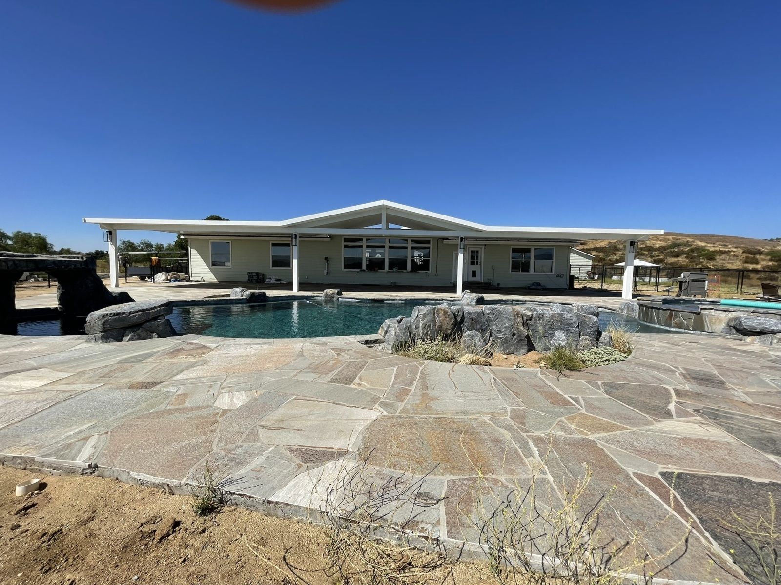Backyard with a swimming pool, stone patio, and a light-colored house under a clear blue sky.
