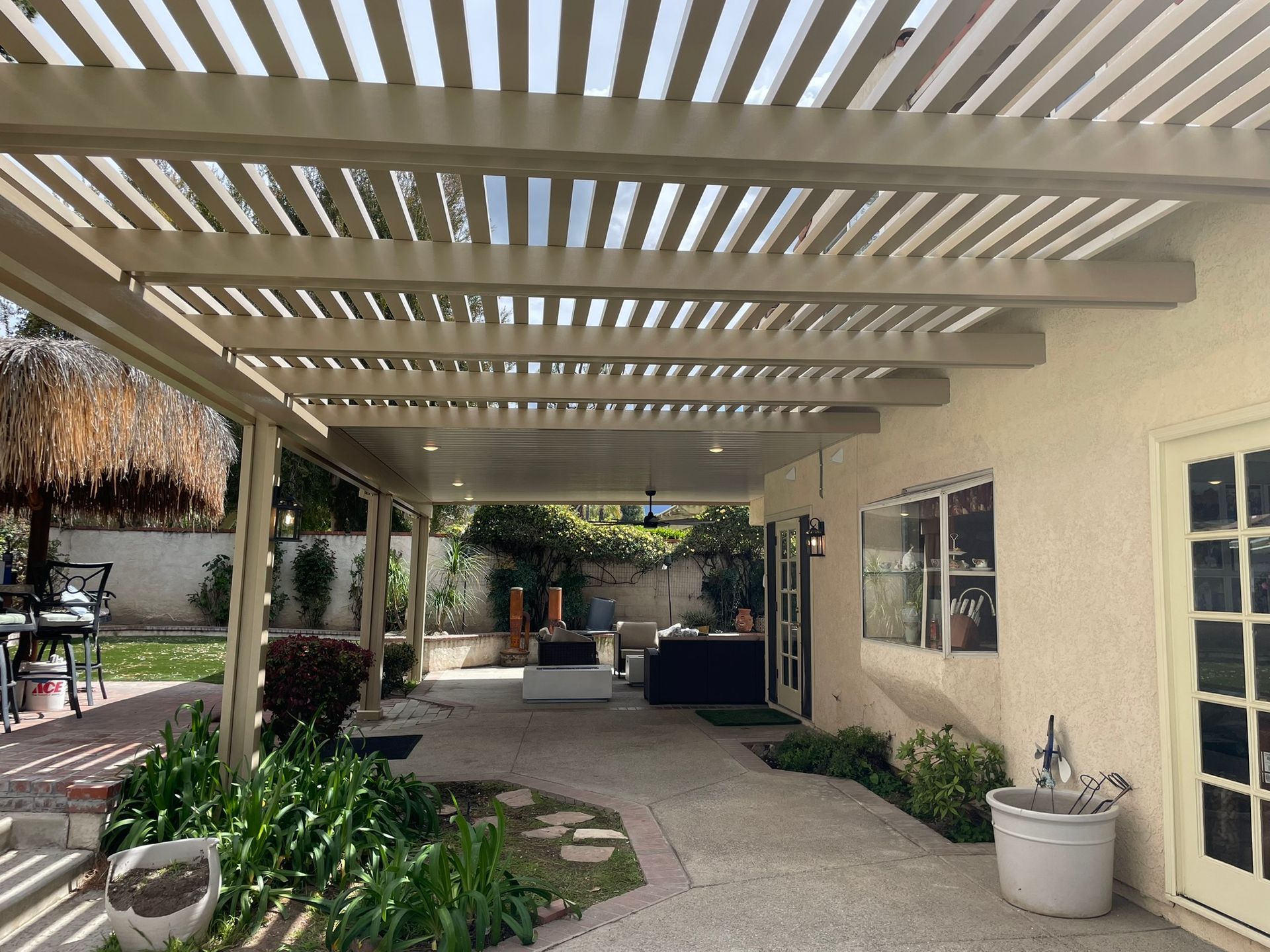 Pergola over a patio, with a view into a backyard. Sunlight streams through the slats.