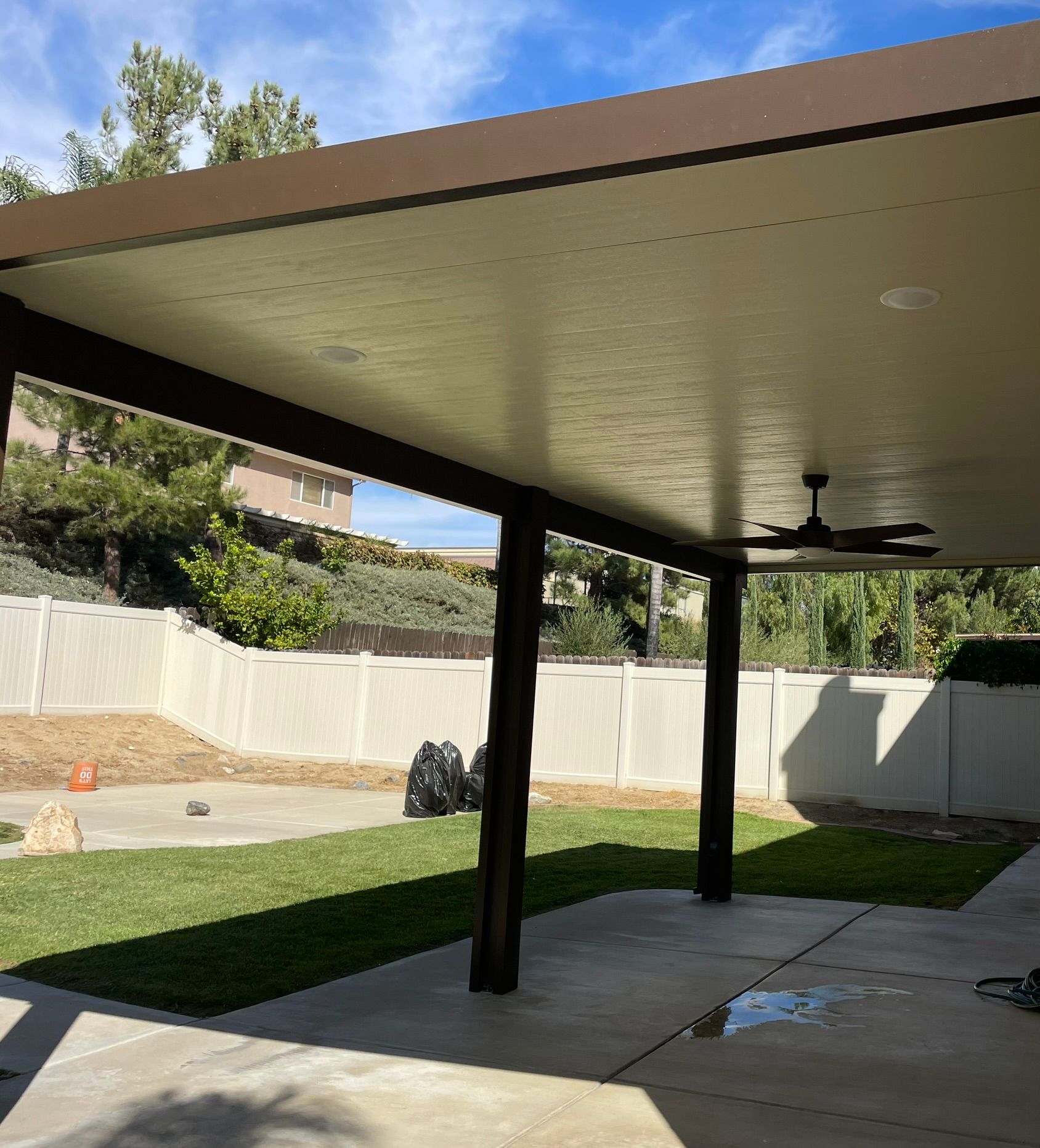 Patio cover with brown beams, white fence, green grass, and blue sky.