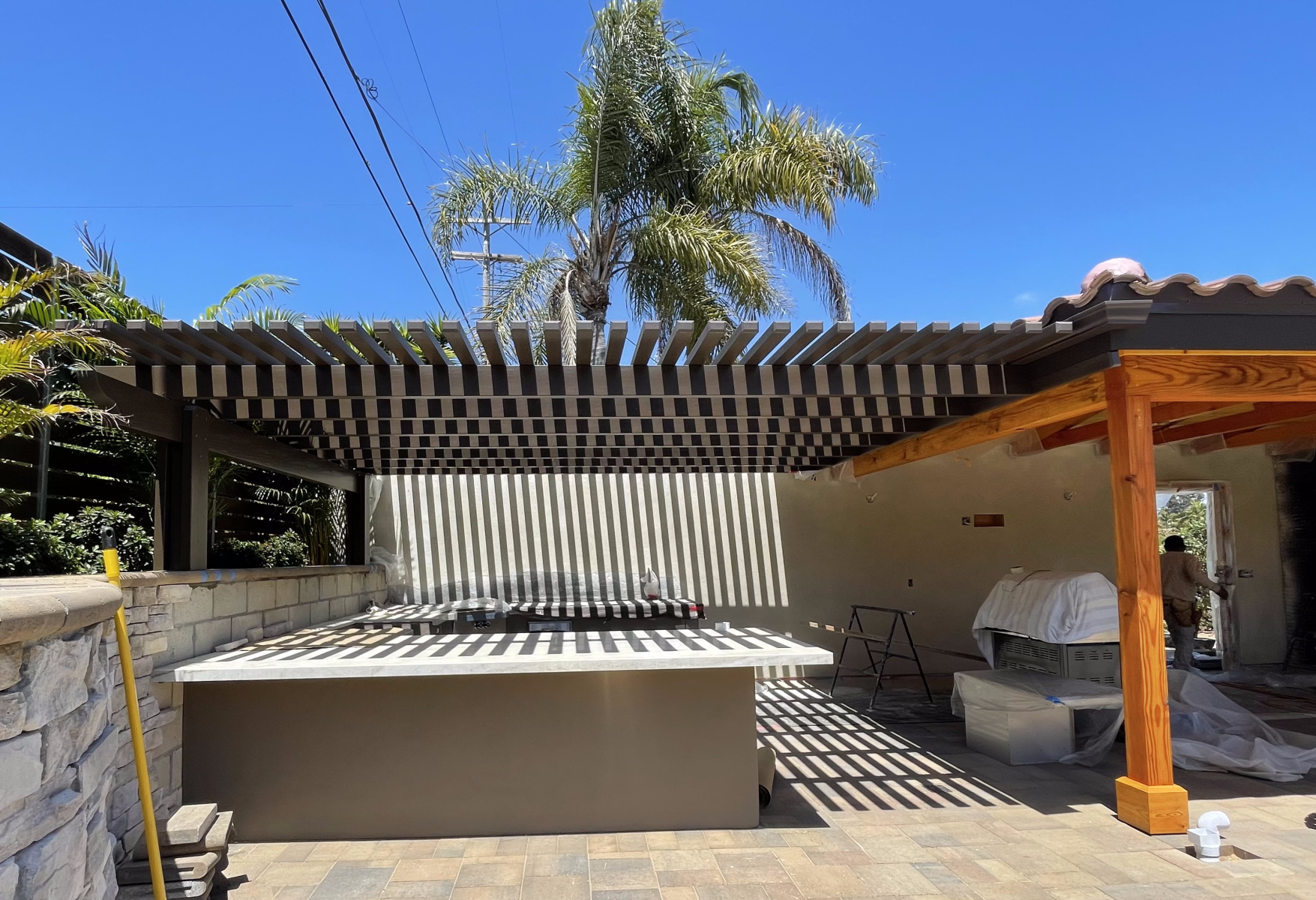 Patio with slatted pergola, outdoor kitchen, and stone wall, casting shadows. Bright sunny day.