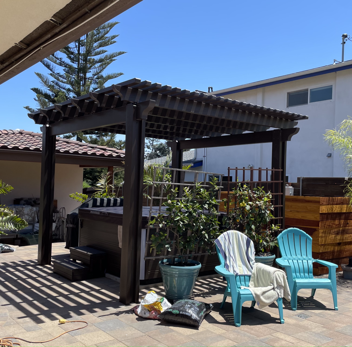 Outdoor seating area with a pergola over a hot tub. Blue chairs, potted plants, and clear sky.