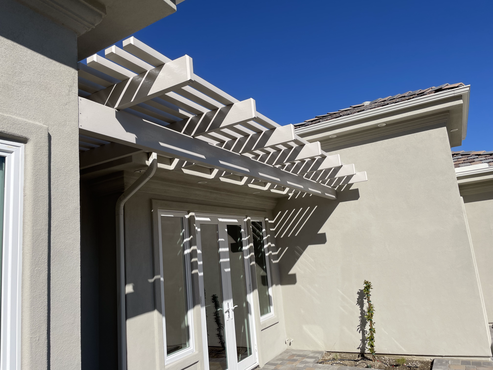 Pergola over French doors, casting shadows on light stucco walls, clear blue sky.