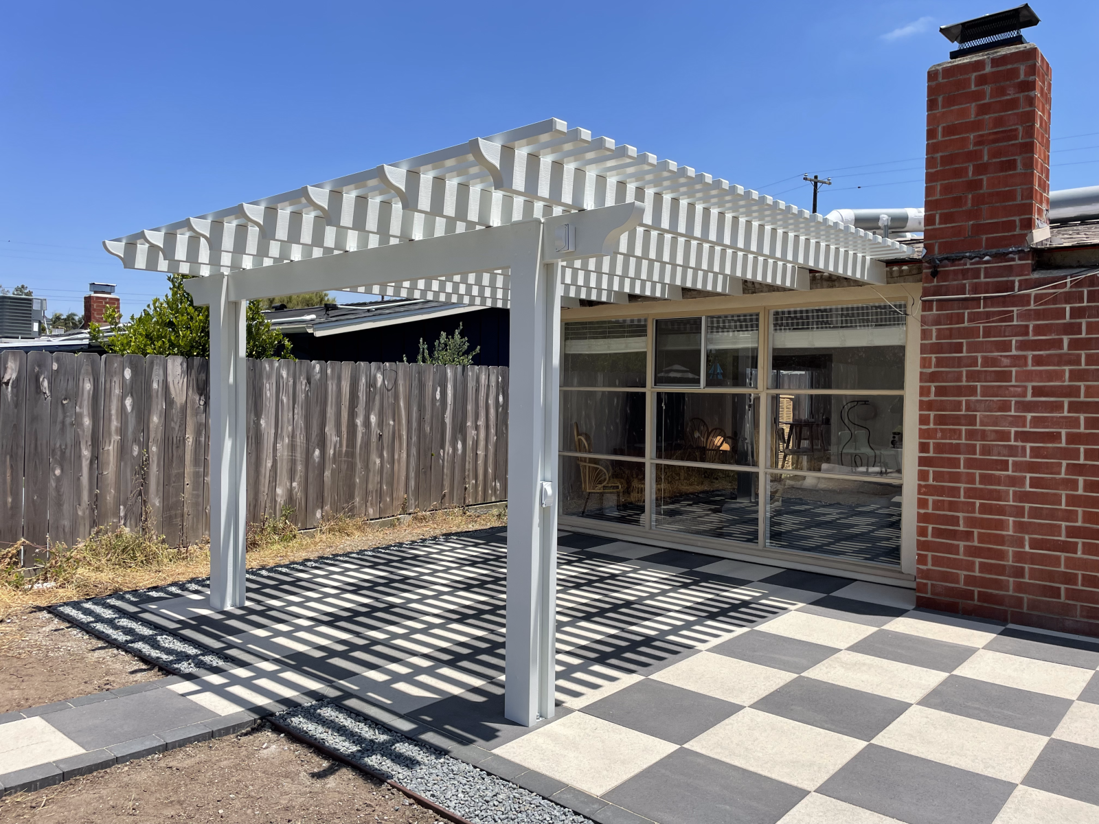 White pergola over a checkered patio, next to a glass-walled structure and a brick chimney.