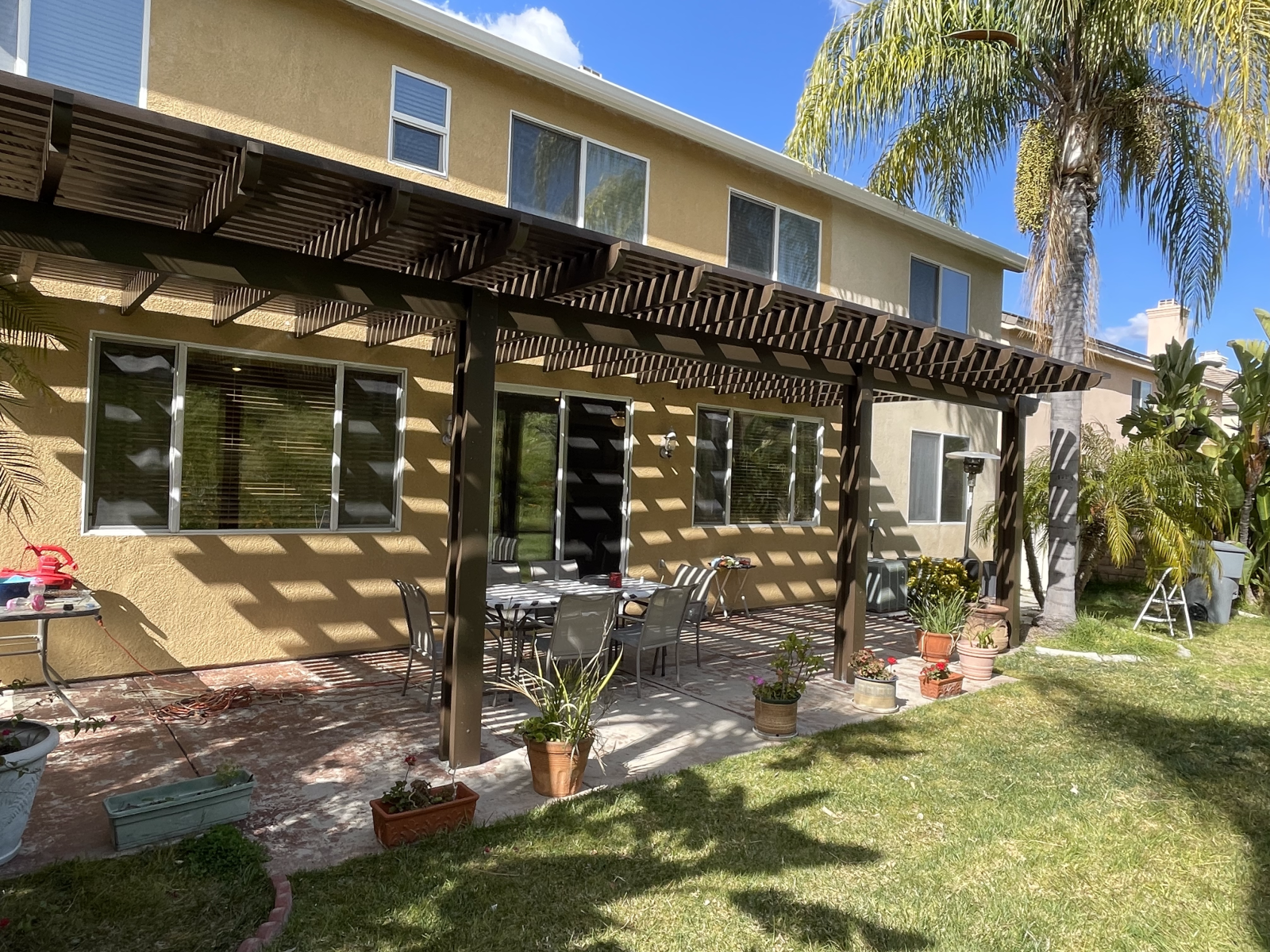 Backyard with a wooden pergola, tan house, and patio furniture. Green grass and a palm tree are visible.