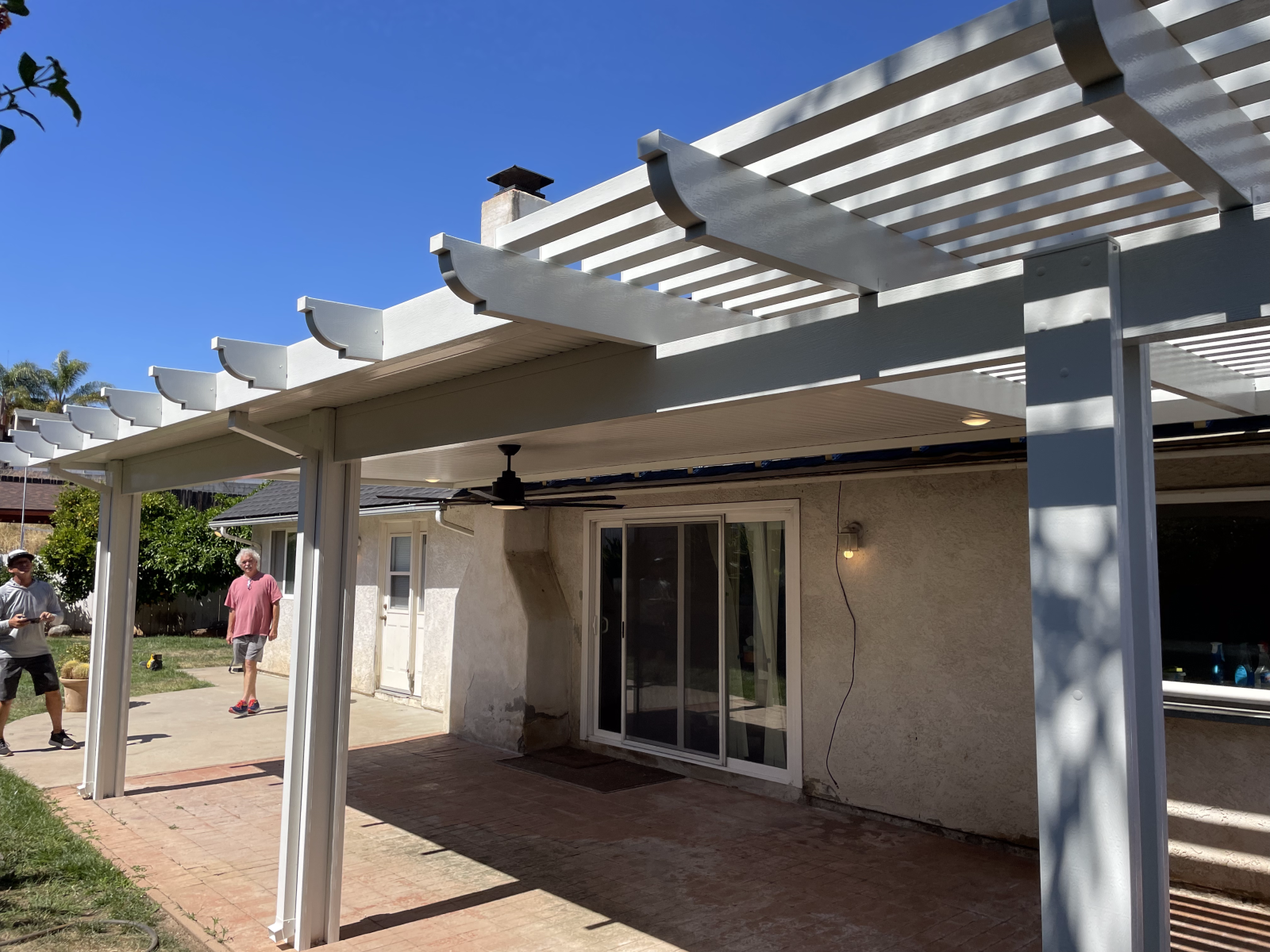 White pergola over a patio with a sliding glass door. Two people stand in the yard.