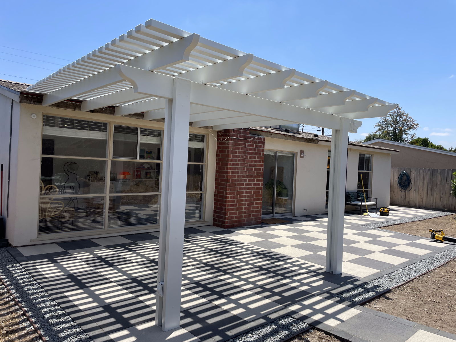 White pergola over a patio with black and gray square tiles. Brick chimney and sliding glass doors visible.