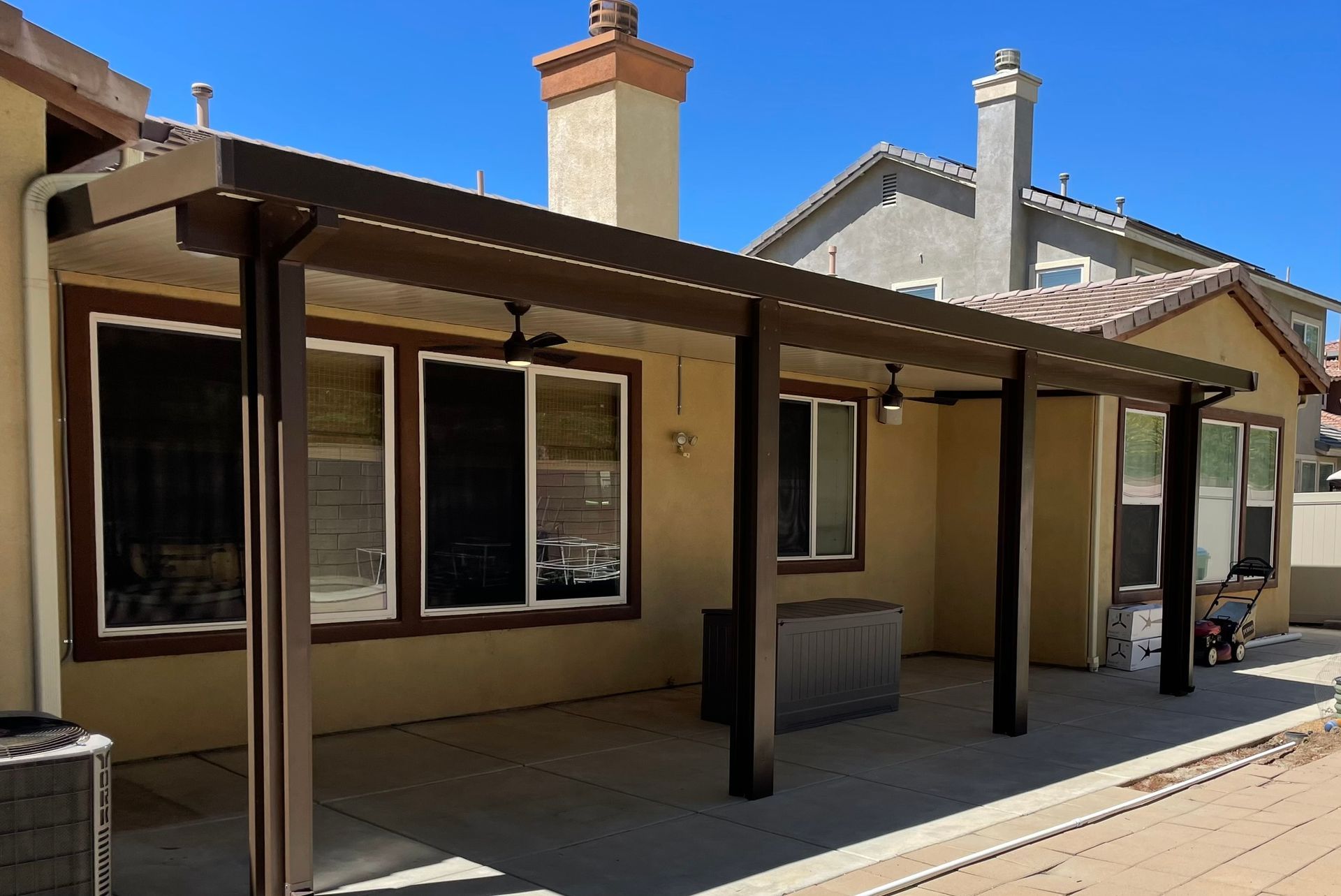 Brown patio cover attached to a beige house, with concrete flooring and a view of other houses.
