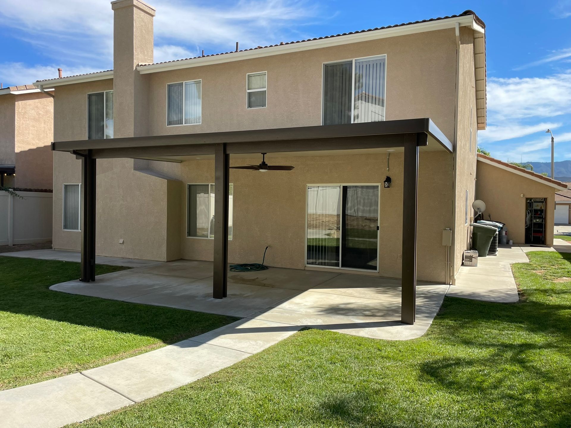 Beige two-story house with a brown patio cover, concrete patio, and green lawn.