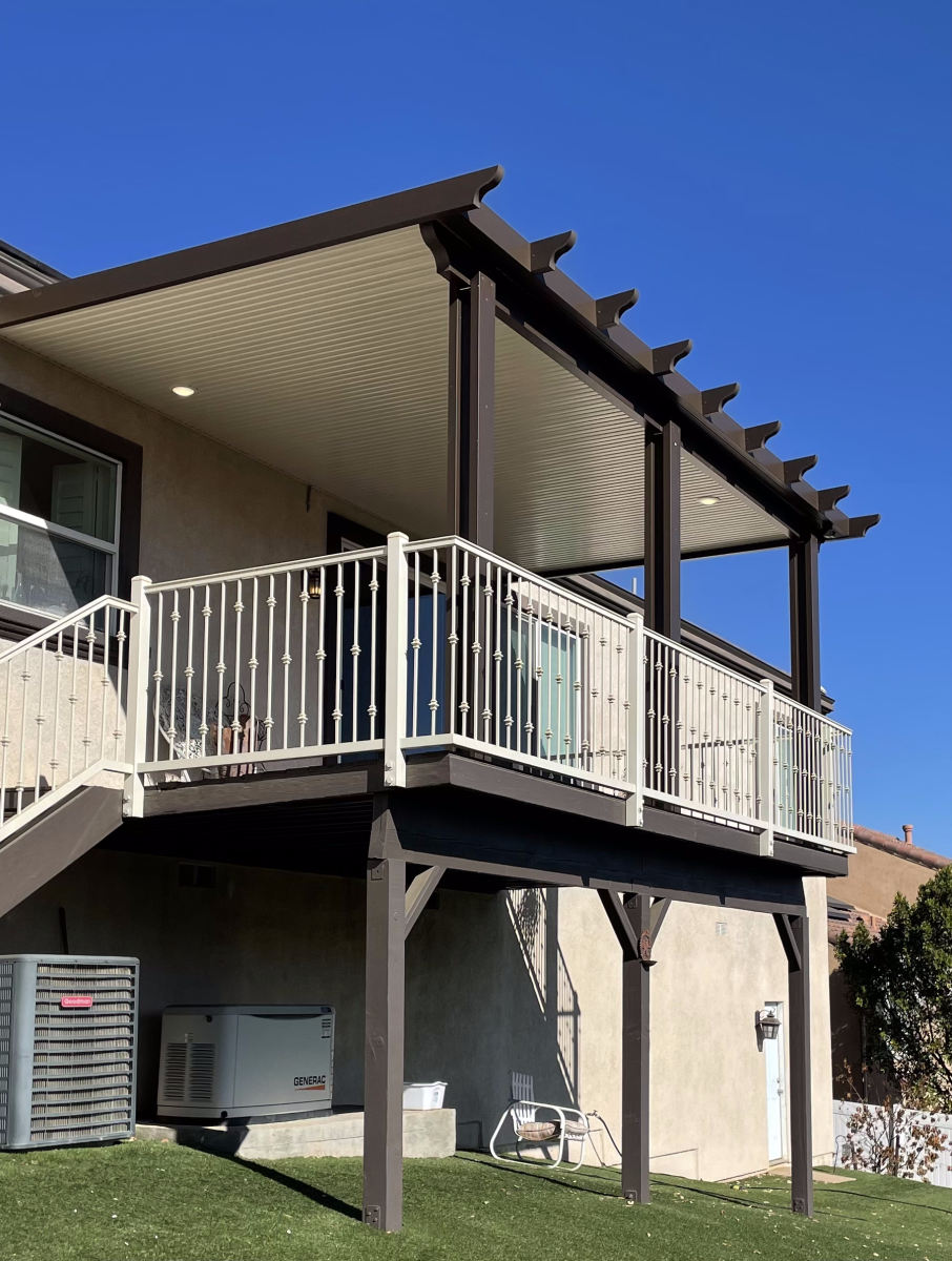 A two-story deck with a brown pergola and white railing, attached to a beige house, under a clear blue sky.