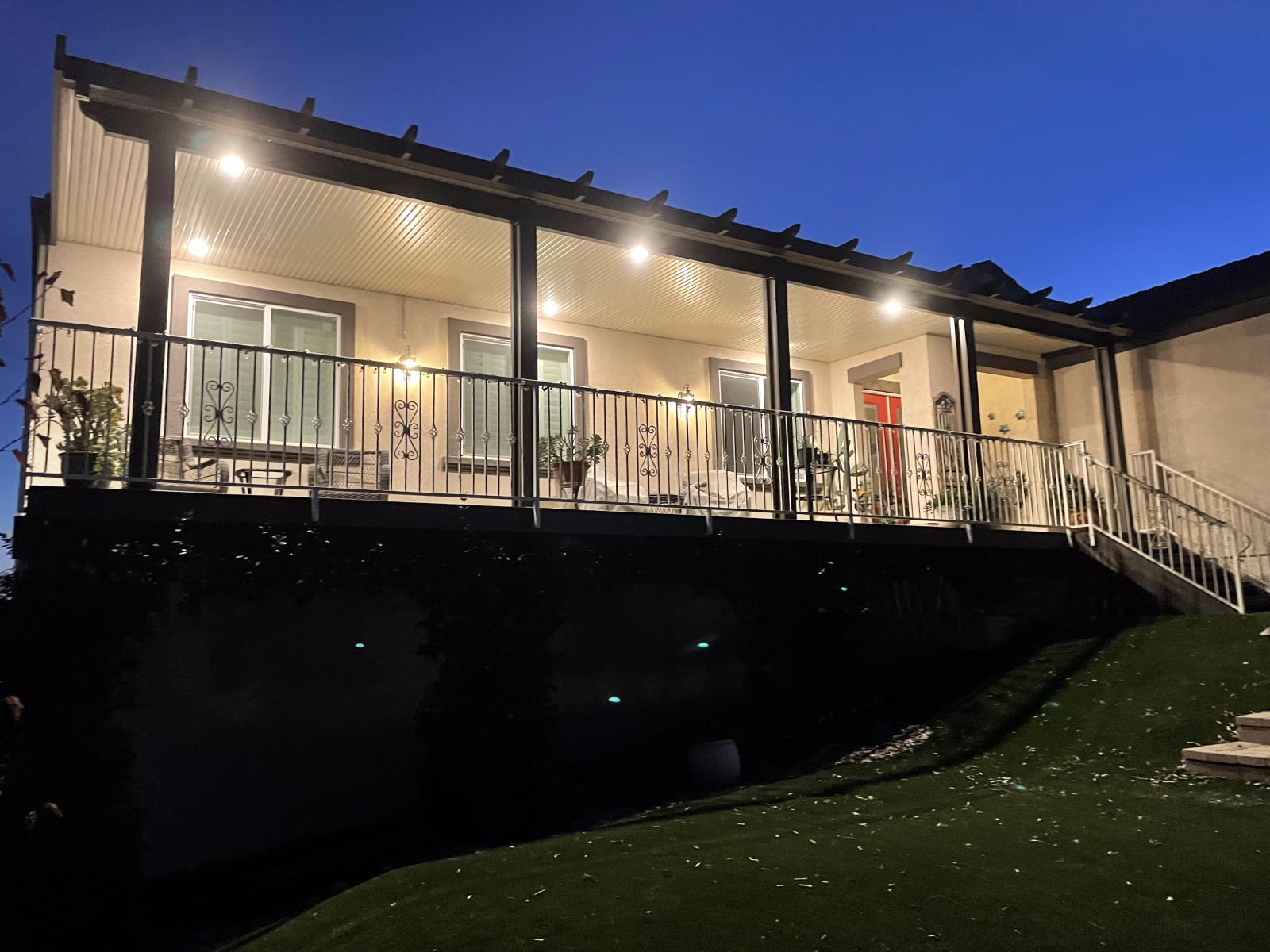 Back porch at dusk with lights on, railing, and stairs leading down to a lawn.