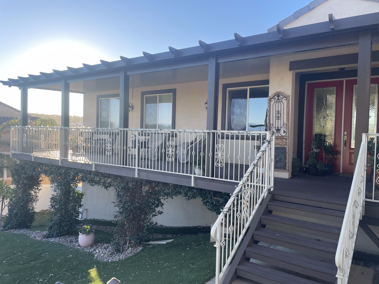Beige house with porch, white railing, dark steps, pergola. Vines grow on the porch supports.